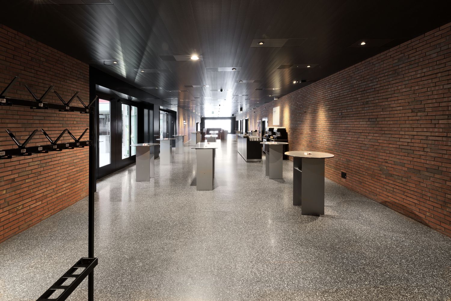 A long foyer between brick walls, a row of grey standing tables with candles on the terrazzo floor, the cloakroom with number
