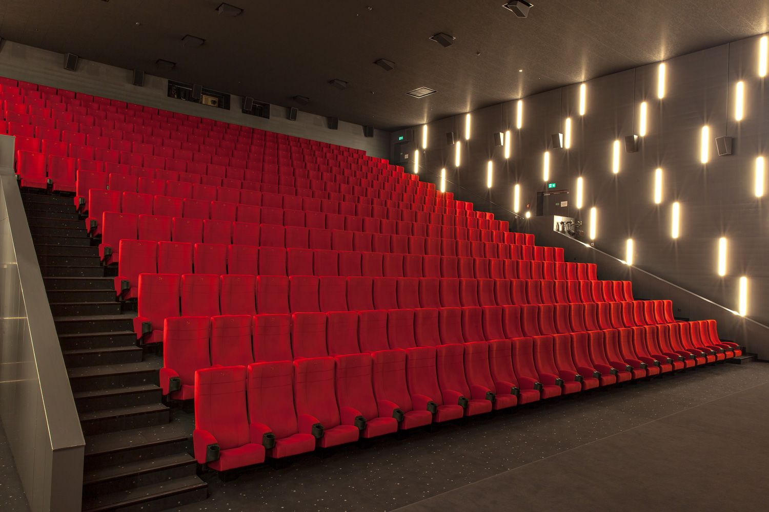 Wide, steeply rising rows of bright red upholstered seats, the stairs with handrail on the left, the grey wall with vertical
