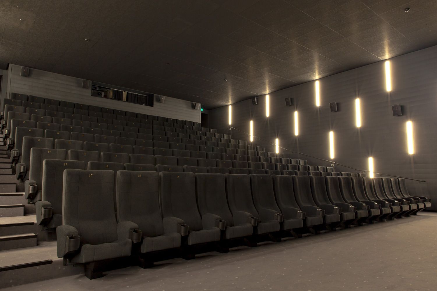 Rising rows of grey-green upholstered seats with gold row numbers, the wall with vertical lights on the right.