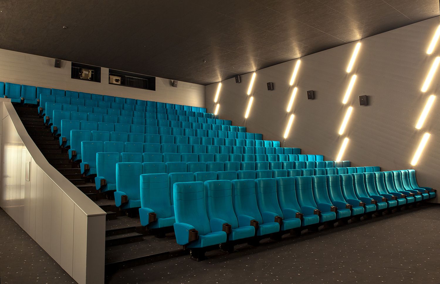 Rising rows of turquoise upholstered seats, with the white balustrade of the stairs on the left.